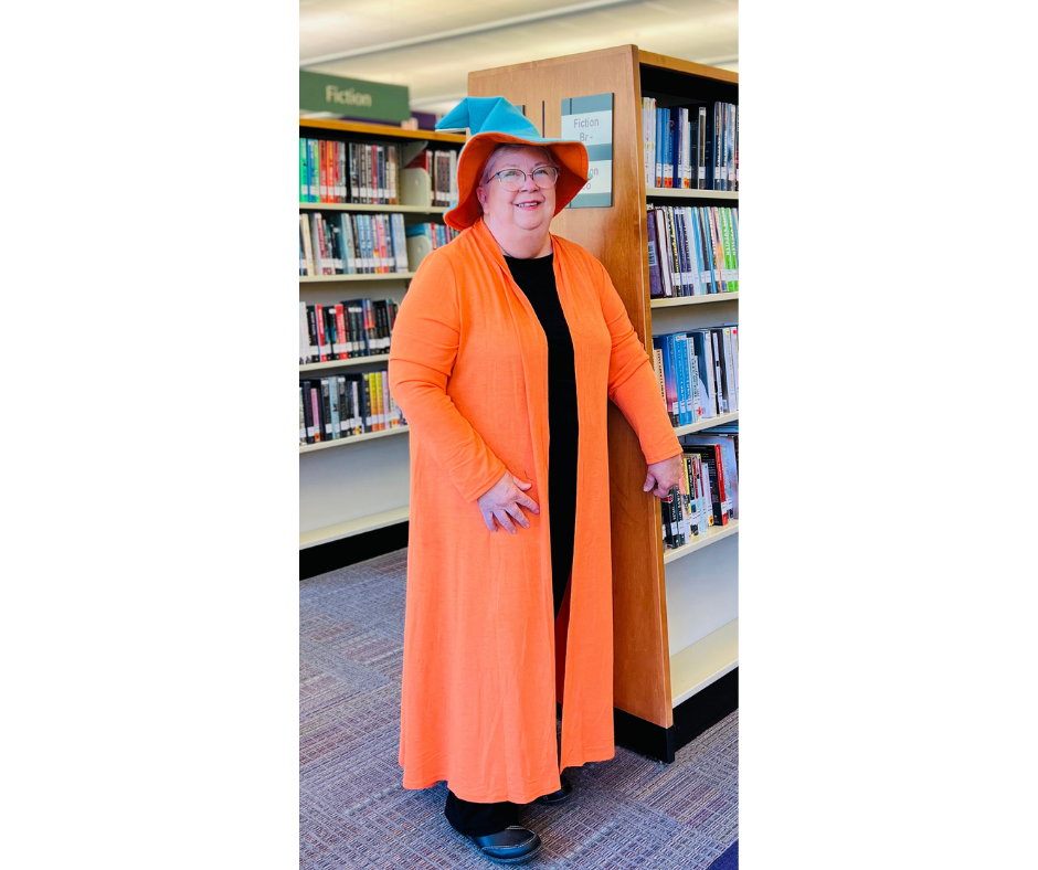 mary The Word Wizard is standing tall in her Orange and Teal Wizard's hat and Orange robe. The rest of her outfit is black. She is in a room full of books on shelves.